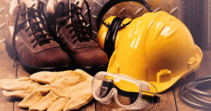 Equipamentos de Proteção Individual (EPI) essenciais para a segurança do trabalho, incluindo botinas de couro, capacete amarelo, abafador de ruídos, luvas de raspa e óculos de proteção, organizados sobre uma superfície de madeira.